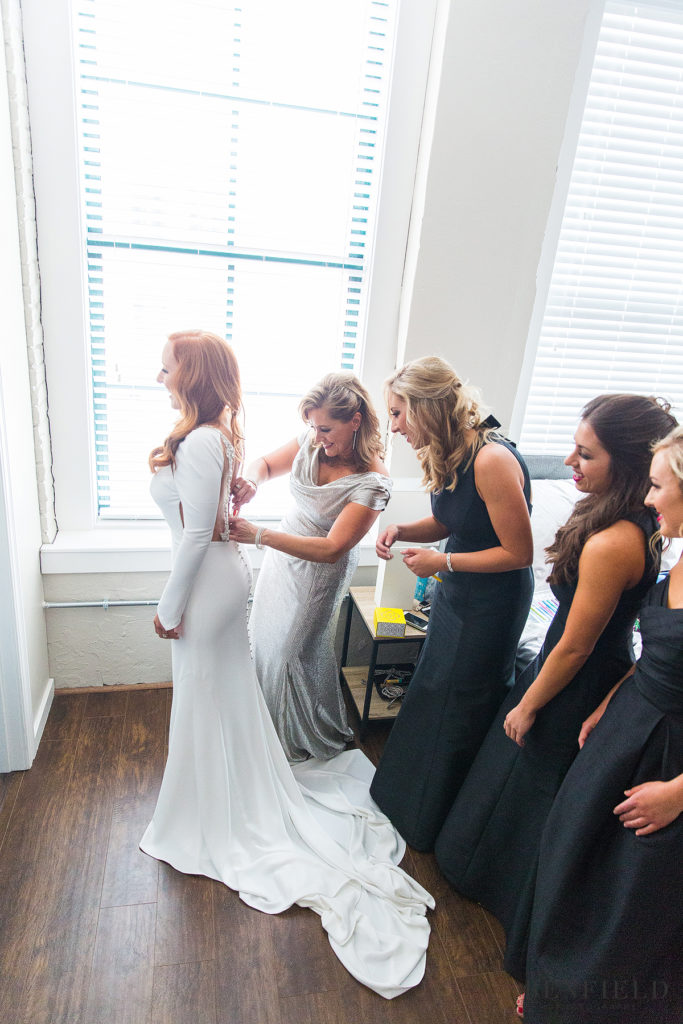 mother of the bride helping the bride into her fitted white dress while the bride's sisters watch on