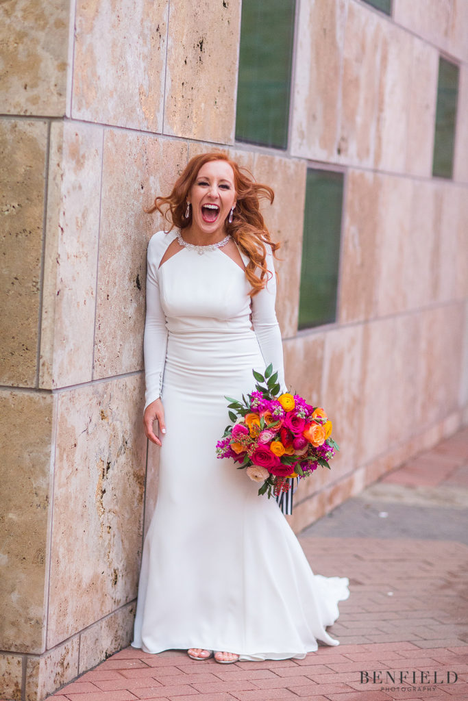 a laughing bride with red hair and a super colorful bridal bouquet wearing a long fitted white dress