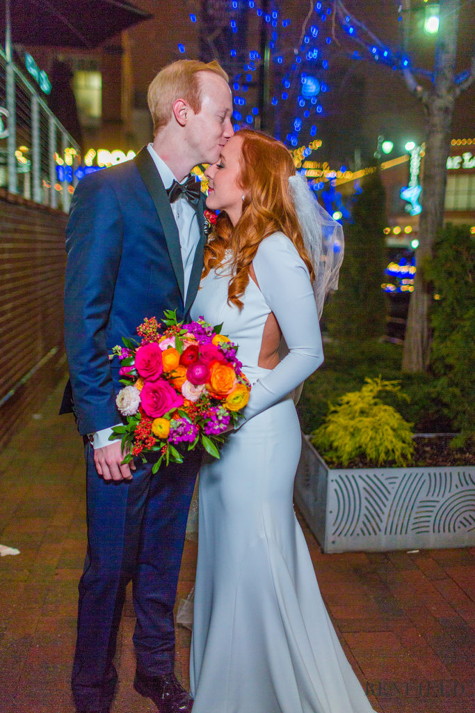 the groom in a blue tux kisses his bride on the forehead at night with blue christmas lights in the background in the power and light district of kansas city (wedding ceremony was at the Gallery Event Space in Kansas City).