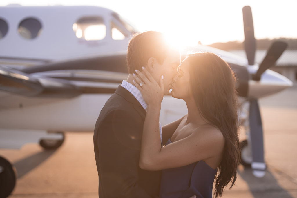 engaged couple kissing in front of an airplane at sunset