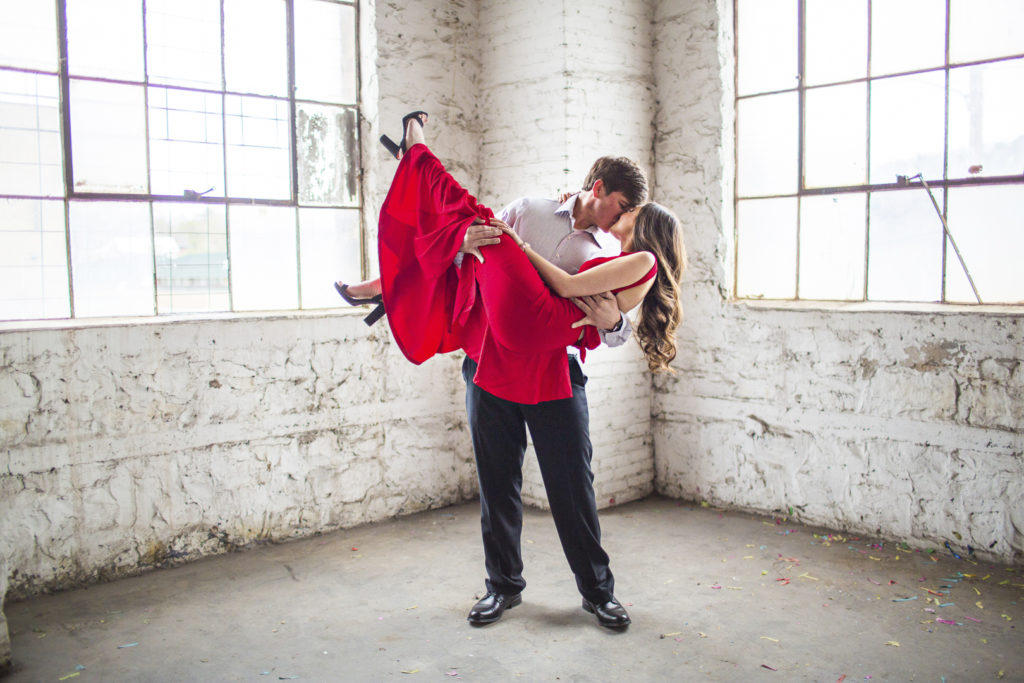 Groom dipping bride during engagement portraits. She is wearing a vibrant red dress in a white parking garage.