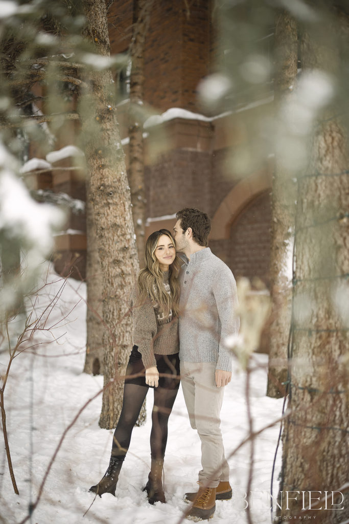 Amanda Stanton's Engagement Photos to Michael took place outside the St. Regis in Aspen. The groom is kissing the bride on her temple surrounded by trees and snowy leaves.