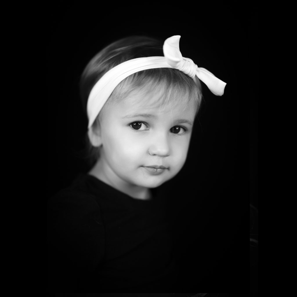 baby two year old portrait in black and white. she is looking at the camera, not smiling, wearing a white bow