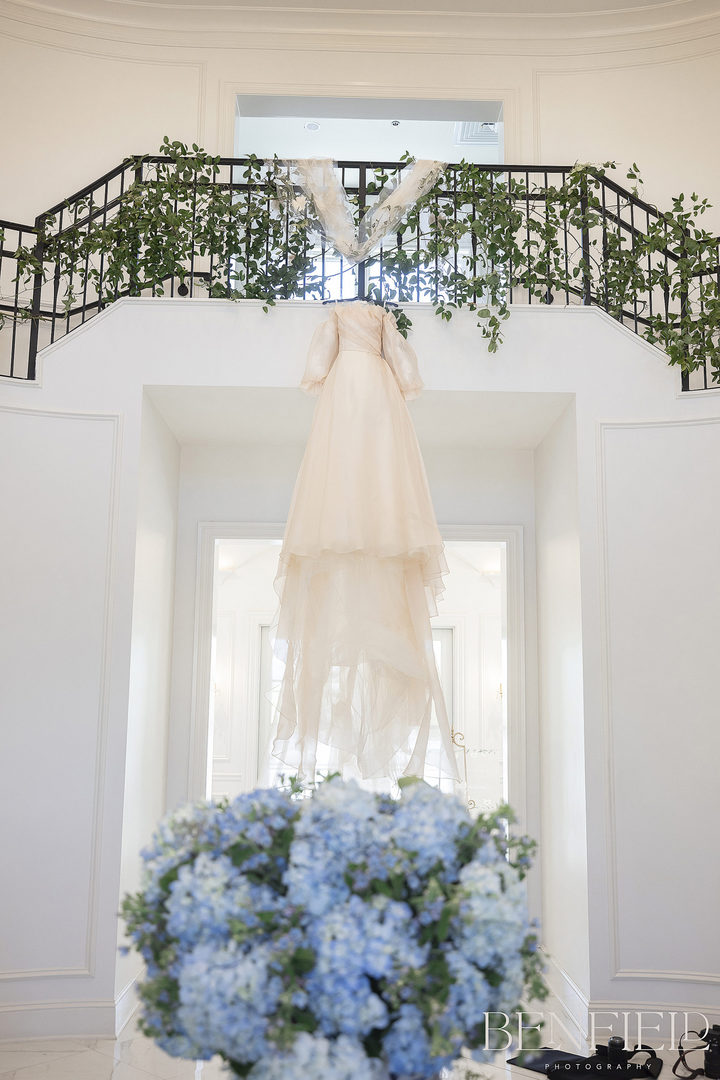 designer luxury blush wedding dress hanging from a grand staircase at Hillside Estate in Crossroads TX under a huge chandelier.