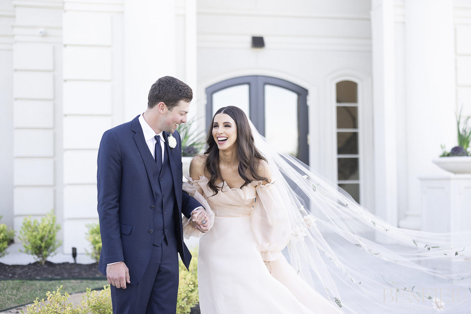 couple laughing outside The Hillside Estate Wedding venue in DFW. The bride's veil is blowing in the wind and you can see the estate behind them.