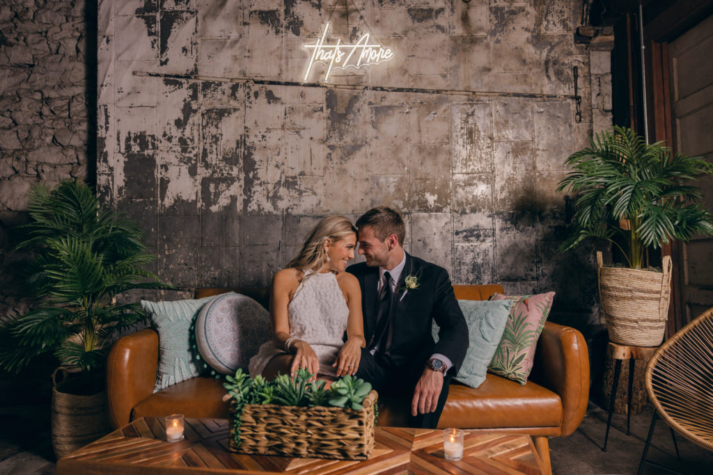 by hiring a photographer who understands lighting you can capture a shot like this of the bride and groom under a neon sign while sitting on a couch during their reception.