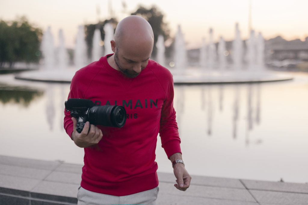 Dale Benfield gives a Canon R5C review and stands in front of a reflective fountain at Kansas City Union Station and Crown Center while wearing a red Balmain sweatshirt and white pants.