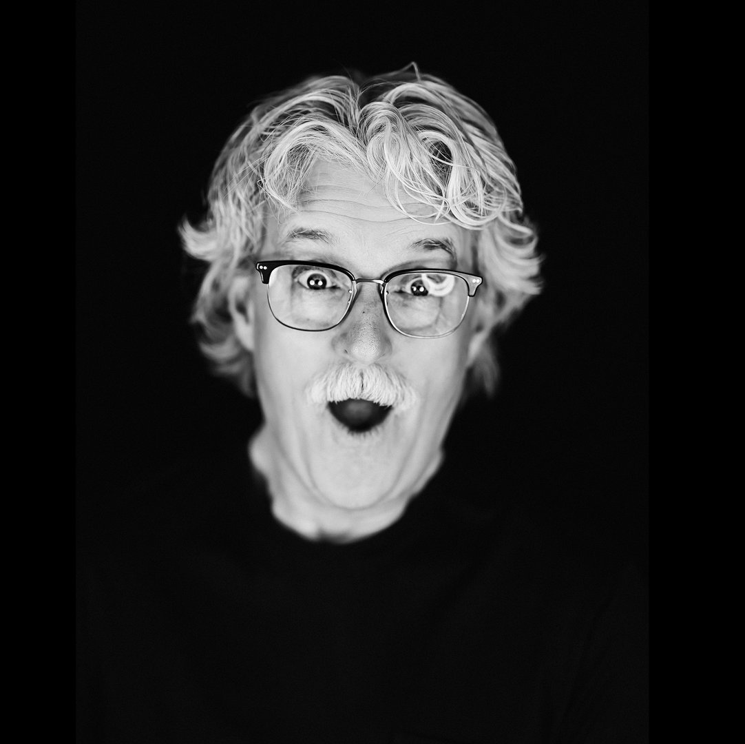 Fine art parent portraits of an aging dad with white curly hair and glasses making a funny face in black and white in front of a black backdrop.