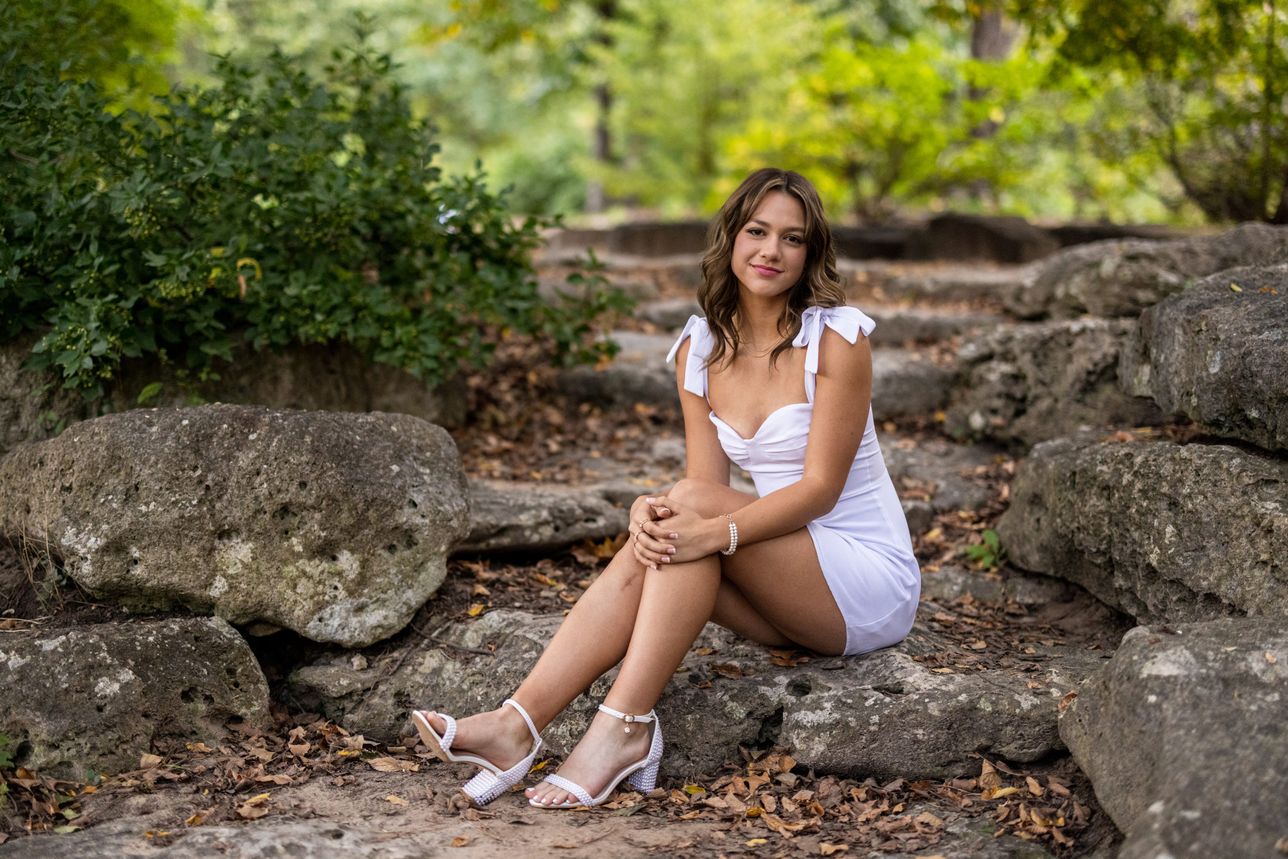 senior pictures in Tulsa at woodward park of a brunette senior in a mini short white dress sitting on a rock staircase.