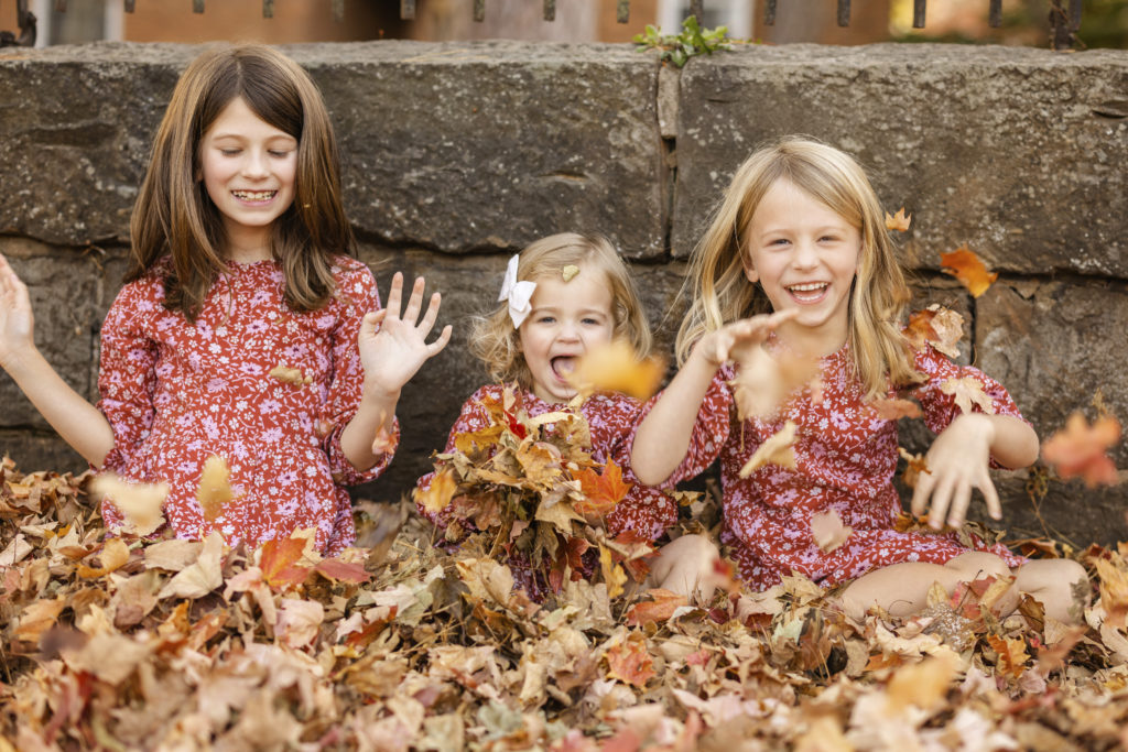 Fall Mini Session in NWA for George Family with 3 little girls in matching floral dresses playing with fall leaves.