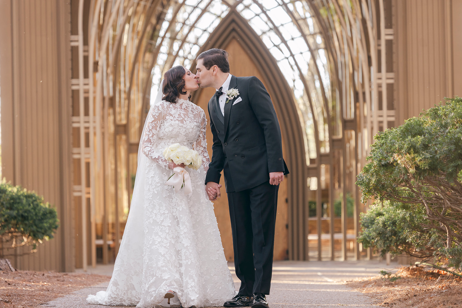 Bride and groom kissing in front of Mildred B. Cooper Chapel in Bella Vista Arkansas, holding bouquet during elegant outdoor wedding portrait